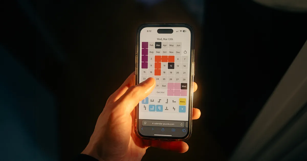 A hand holding a phone playing The Calendar Puzzle in the browser: the Wed, Mar 12th board with coloured pieces placed and the shape bank below